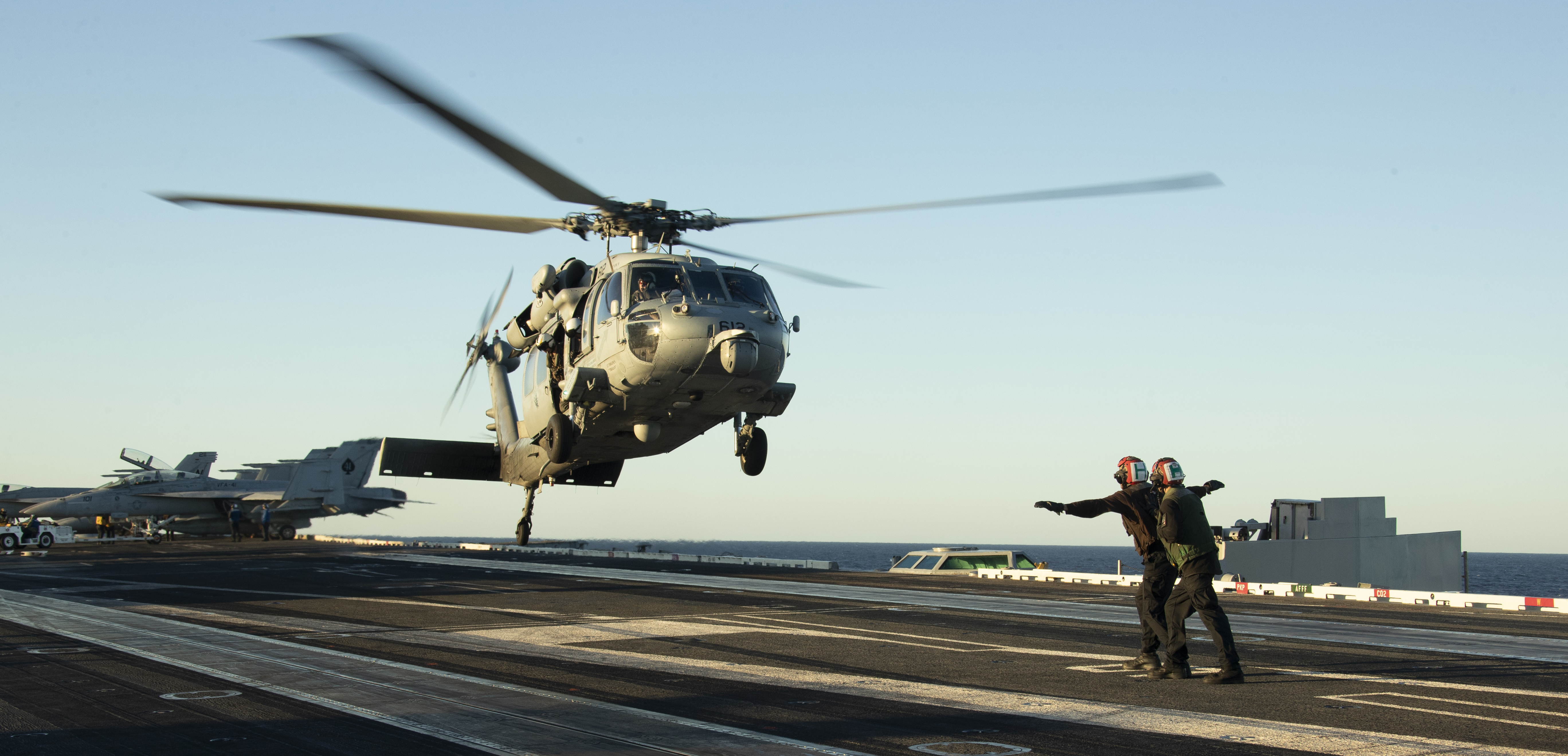Two landing signal officers guide a helicopter landing on an aircraft carrier