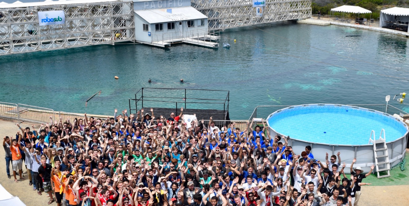 A large group of attendees is in front of a large body of water, a smaller pool is on the right